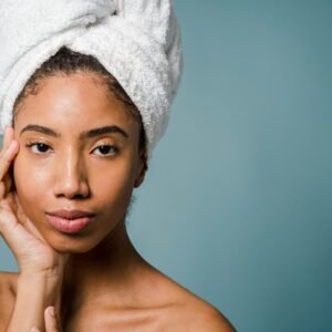 Tranquil young black female with pure skin and white towel on head touching face and looking at camera after spa procedures against blue background